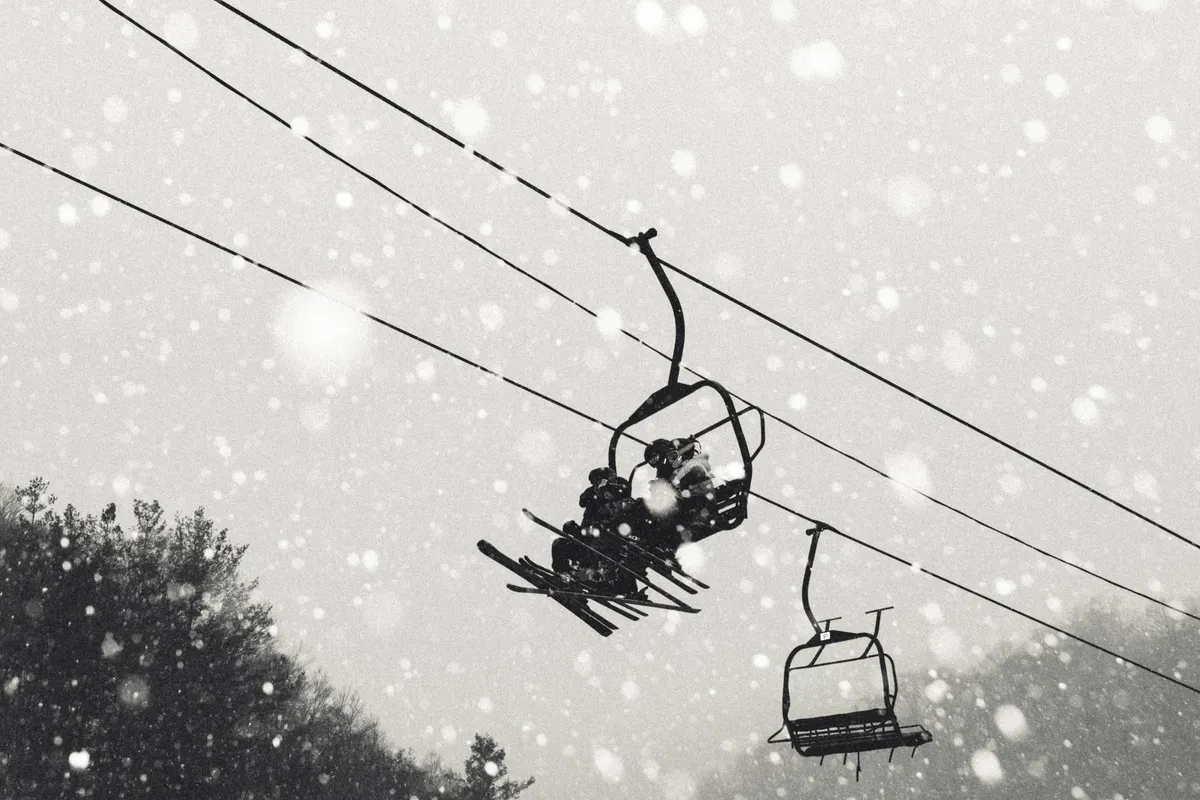 This image captures an exterior winter scene with ski lift chairs ascending through gently falling snow, framed by snow-covered trees in the background. The quiet, serene atmosphere emphasizes the beauty and tranquility of mountain life, inviting viewers to imagine themselves exploring or relaxing in peaceful solitude.