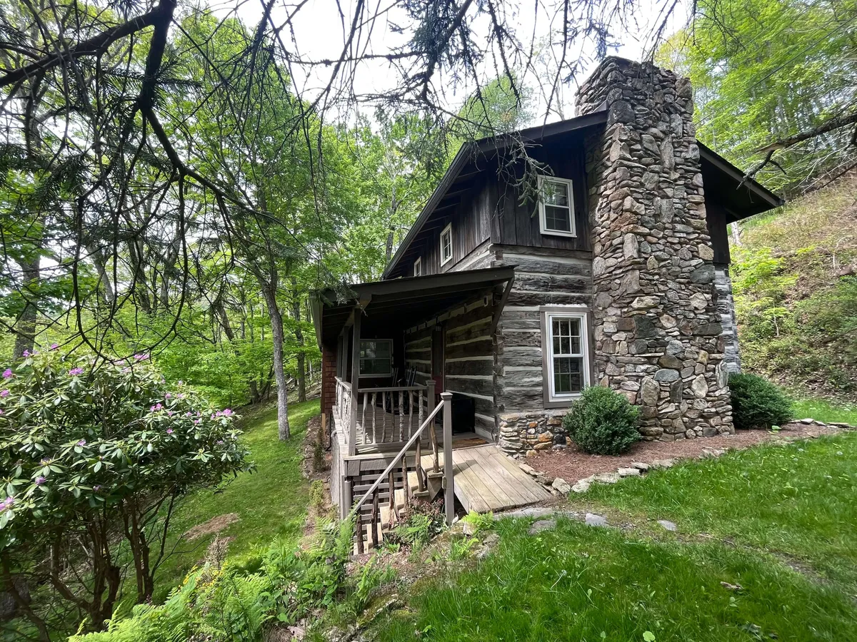 The image showcases a classic, hand-crafted log cabin nestled among lush greenery and flowering shrubs in the Appalachian Mountains. The inviting wooden steps lead up to a rustic porch, complete with a beautiful stone chimney, all set within a peaceful, gated resort community.