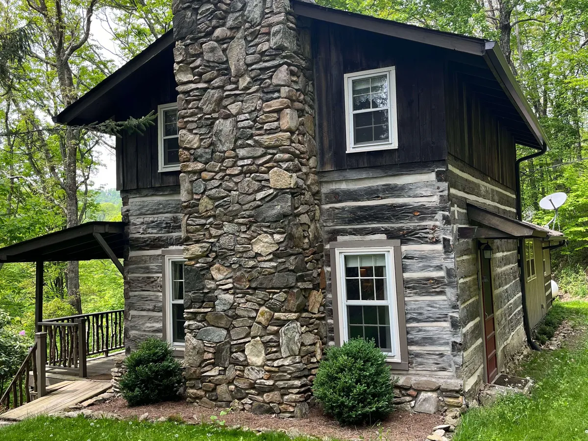 A classic cabin exterior with sturdy stone chimney, log walls, and a wooden porch nestled among lush greenery. The porch is well-positioned for sipping morning coffee and wildlife watching, echoing the wooded yard and porch features mentioned in the section.