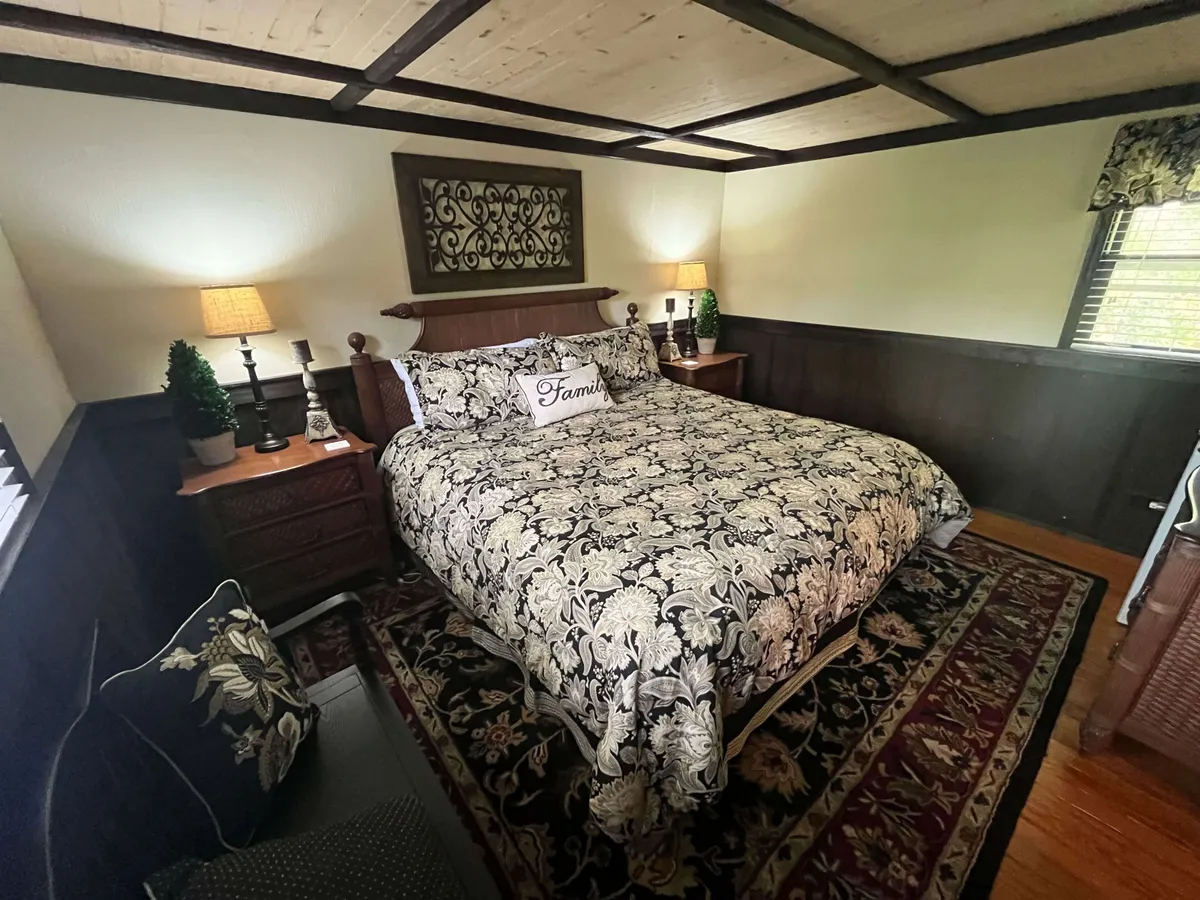 A warmly lit bedroom with an ornate bed, plush bedding, nightstands with lamps, decorative wall art, and a cushy chair in the corner. The wooden accents and layered textiles create a feeling of comfort and timelessness, inviting relaxation.