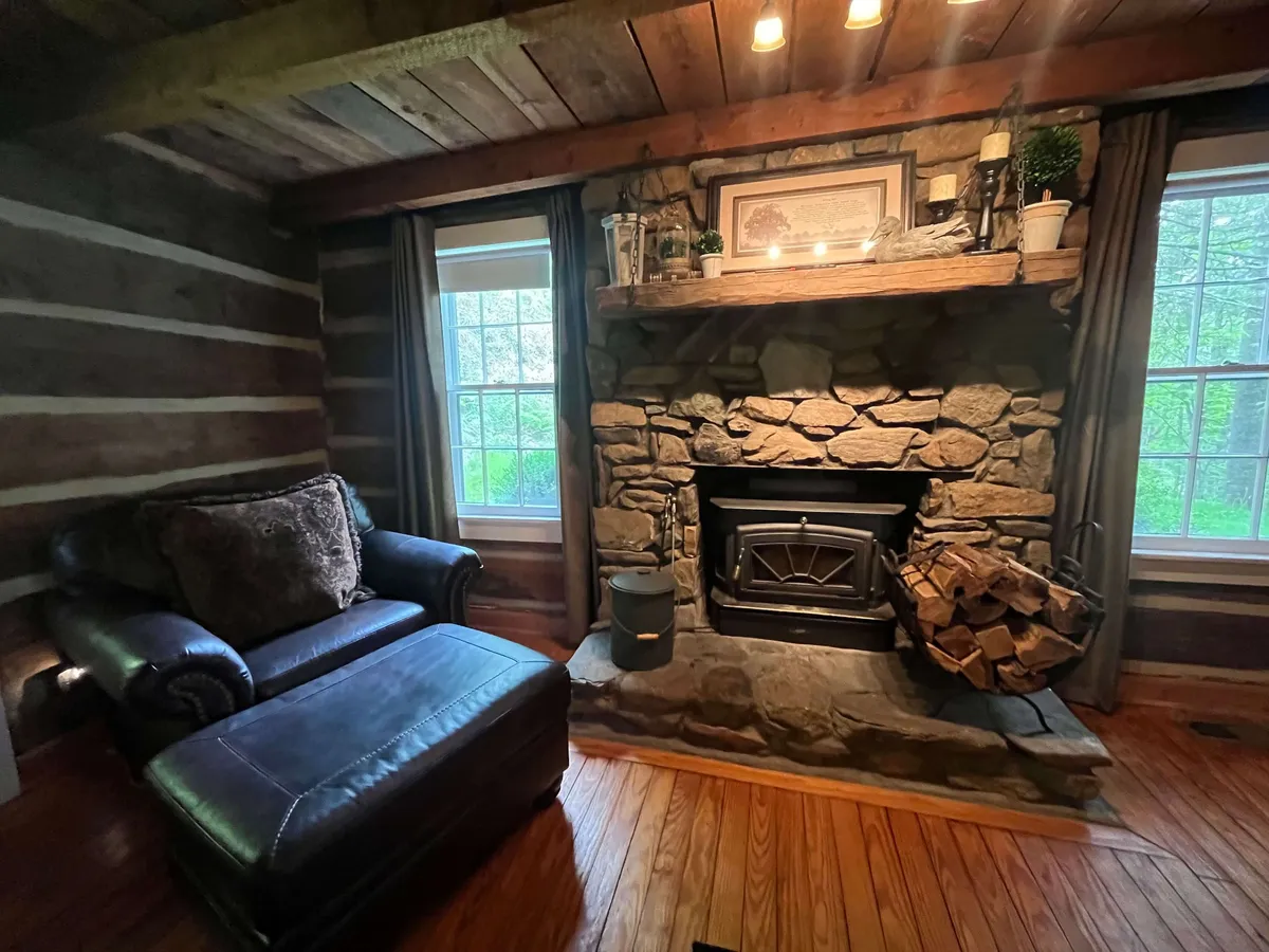 A warmly lit living room featuring a substantial stone fireplace, a leather chair positioned for relaxation, exposed wooden beams overhead, and windows that frame the surrounding woods.