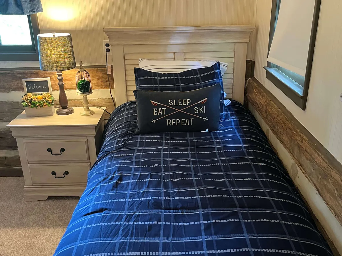 A bedroom with inviting plaid bedding, a decorative pillow with a warm message, and a soft lamp on a nightstand. A welcome sign and scenic window add a restful, intimate touch, perfect for quiet mornings or evenings together.