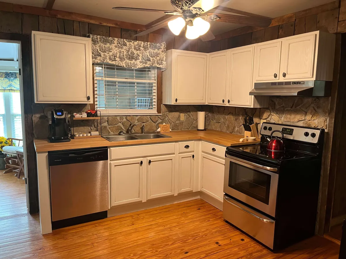 This image showcases a warm, newly renovated kitchen featuring stainless steel appliances, natural wood countertops, a charming stone backsplash, and a kitchen window dressed in light curtains. The scene exudes comfort and functionality, suggesting home-cooked meals and joyful mornings.