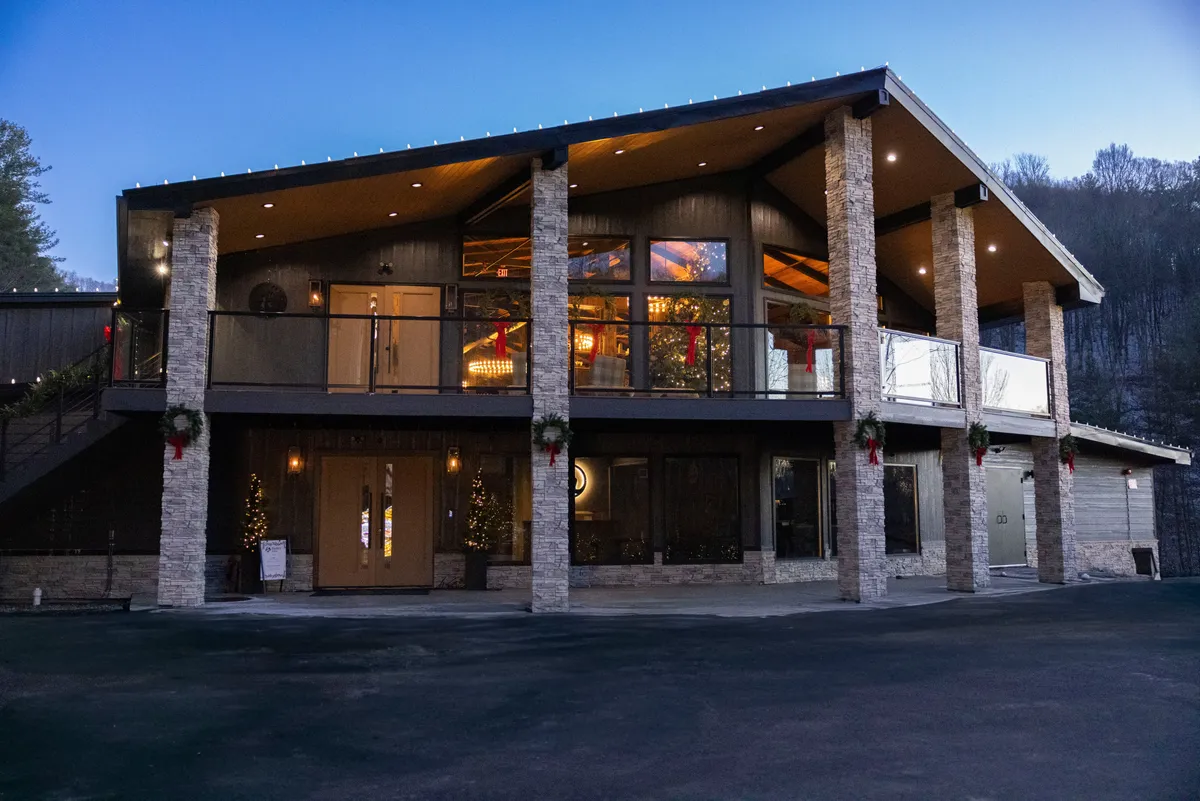 A welcoming entrance with double doors, large windows that reveal tastefully decorated interiors, and a balcony overlooking the landscape. The exterior is framed by greenery and invites you into the cabin, establishing a sense of arrival and connection with the outdoors.