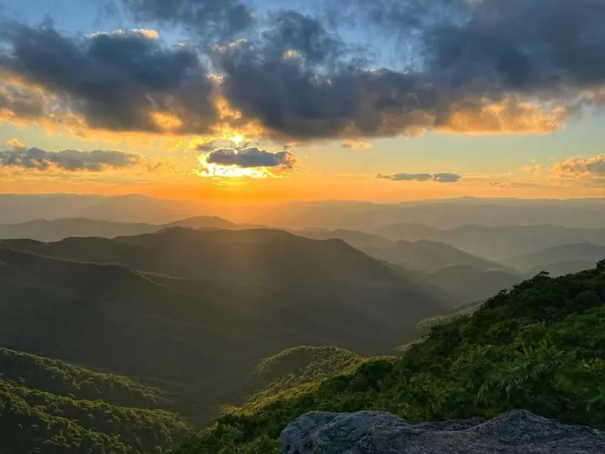 A breathtaking mountain sunset stretches across the horizon, with rolling ridges and a vibrant sky above lush greenery. The scene evokes peace, adventure, and the beauty of the natural world just beyond the front door.