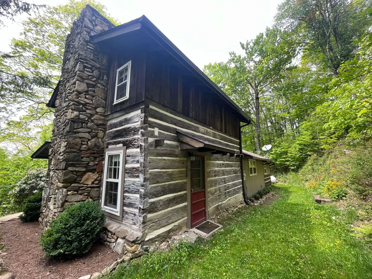This image features a cozy mountain cabin exterior with a prominent stone chimney, rustic wooden logs, a cheerful red door, and lush greenery surrounding the home. The inviting porch area is framed by natural forest, creating a sense of seclusion and warmth.