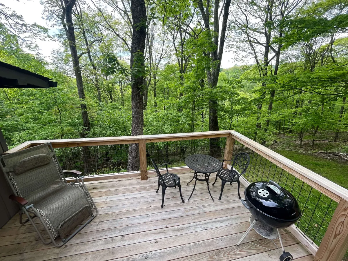 A cozy outdoor seating arrangement with a small round table, two chairs, a grill, a recliner, and tall trees surrounding the space. The area is perfect for relaxing, dining, or watching wildlife, reinforcing the cabin’s seamless transition from inside to outdoor living.