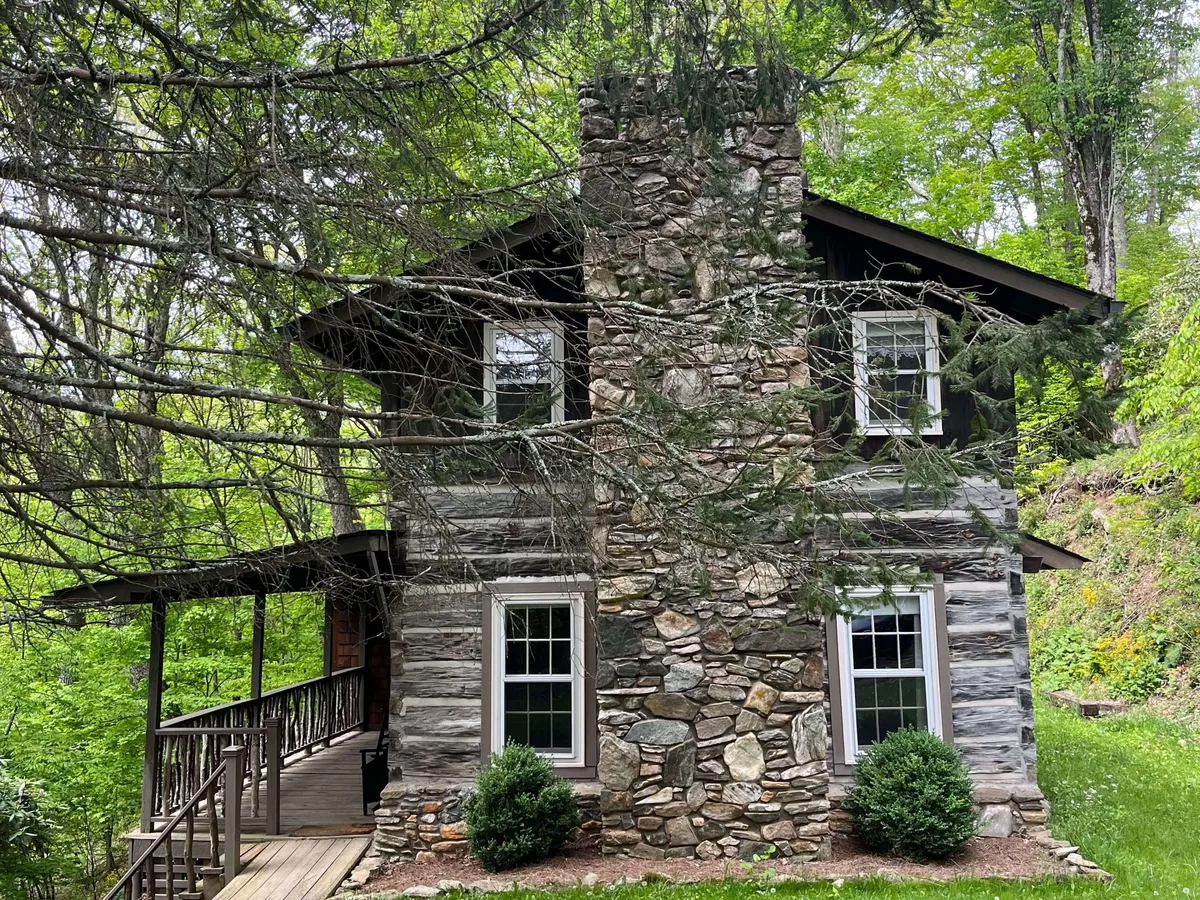 A cozy, rustic cabin exterior with a prominent stone chimney, a classic wooden porch, large windows, and lush greenery all around. The porch looks inviting for relaxing after a day of adventure, emphasizing the close connection between the home and its natural surroundings.