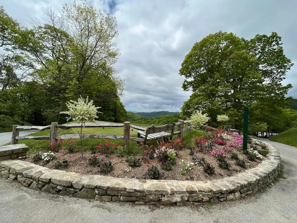 A serene outdoor area with a flower bed, wooden bench, stone wall, and lush surrounding trees, inviting moments of quiet reflection or community gatherings in a historic environment.